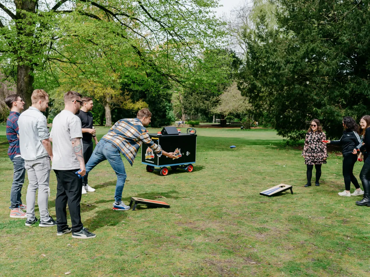 Eine Gruppe spielt Cornhole auf einer Parkwiese, der Battle-Wagen steht dazwischen